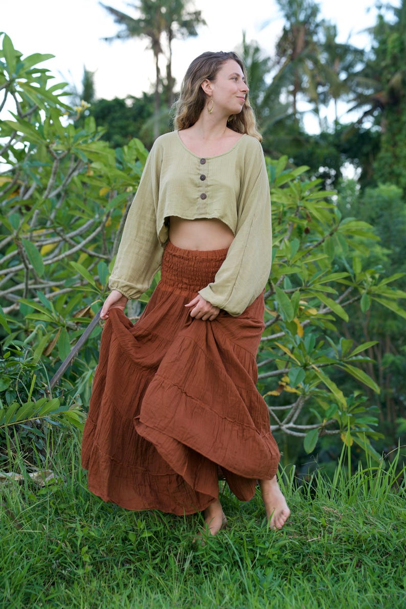 May include: A woman wearing a two-piece outfit. The top is a light green, long-sleeved, cropped blouse with three buttons. The bottom is a long, flowing, rust-colored skirt. The outfit is worn outdoors in a natural setting.