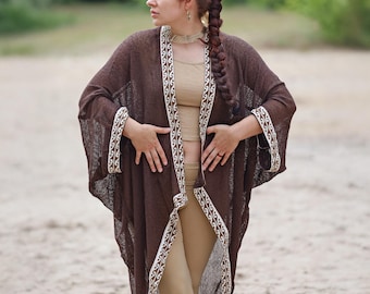 Può includere: Un cardigan marrone, stile kimono, con apertura frontale e rifiniture con motivi bianchi e beige. Il cardigan è indossato sopra un top e pantaloni beige. L'immagine è scattata all'aperto su una spiaggia sabbiosa.