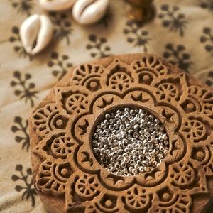 May include: A wooden block with an intricate carved mandala design. The center of the mandala is a circular depression filled with silver beads.