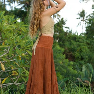 May include: A woman wearing a light green halter top and a long, tiered rust-colored skirt. The skirt has a smocked waistband. The background features lush green foliage and palm trees, suggesting a tropical setting.