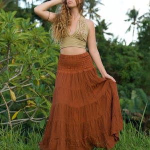 May include: A woman wearing a two-piece outfit outdoors. The top is a light green halter with a geometric design. The skirt is a long, tiered, rust-colored maxi skirt. The background features lush green foliage and palm trees.