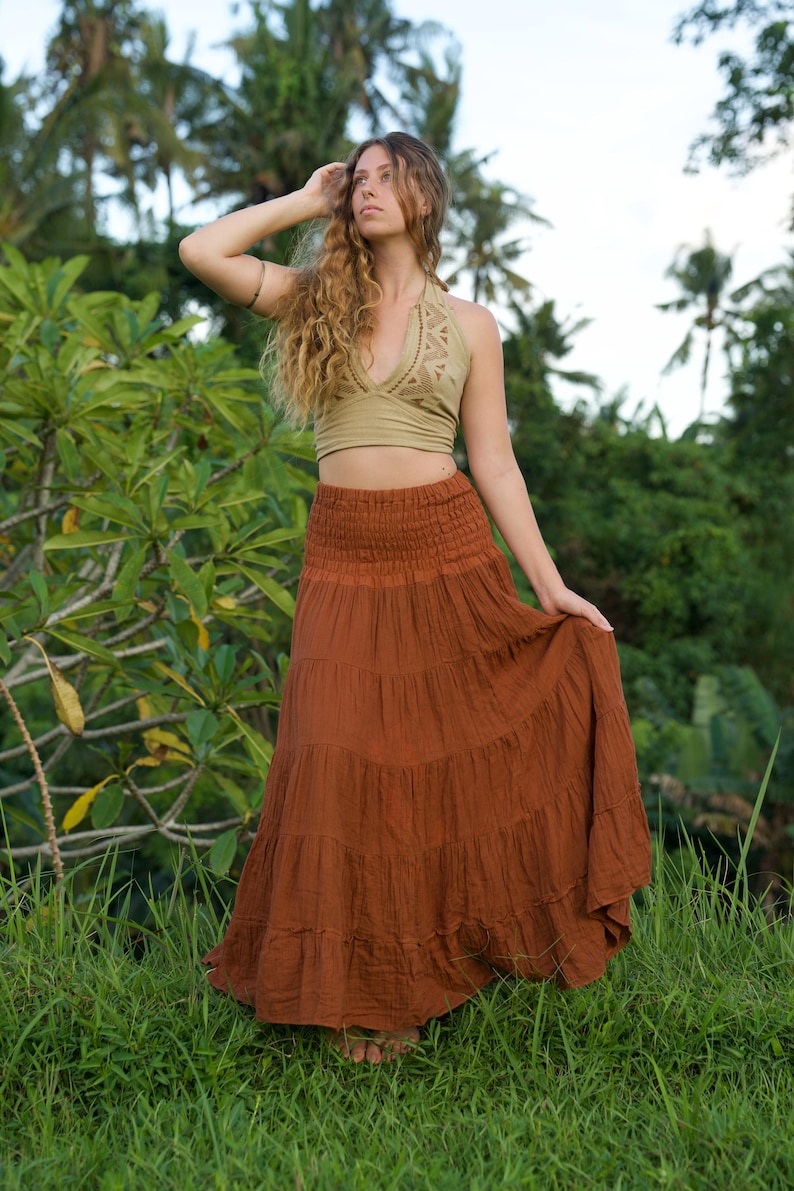 May include: A woman wearing a two-piece outfit outdoors. The top is a light green halter with a geometric design. The skirt is a long, tiered, rust-colored maxi skirt. The background features lush green foliage and palm trees.
