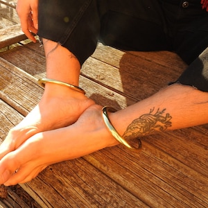 K&ouml;nnte beinhalten: Zwei goldfarbene Metallarmreifen, die an den Kn&ouml;cheln einer Person getragen werden, die auf einer Holzoberfl&auml;che sitzt. Die Person hat ein Herz-Tattoo am Unterschenkel.