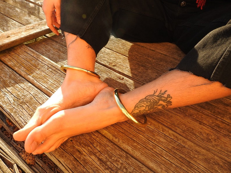 K&ouml;nnte beinhalten: Zwei goldfarbene Metallarmreifen, die an den Kn&ouml;cheln einer Person getragen werden, die auf einer Holzoberfl&auml;che sitzt. Die Person hat ein Herz-Tattoo am Unterschenkel.