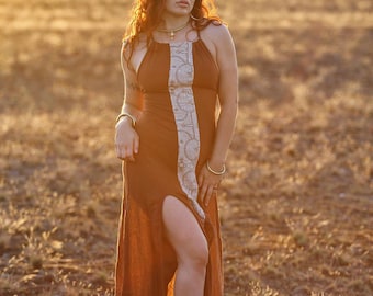 May include: A brown halter dress with a high slit and a decorative white and beige panel down the front. The dress is worn outdoors in a field. The model is wearing gold jewellery, including a necklace and bracelets. The dress is floor length.