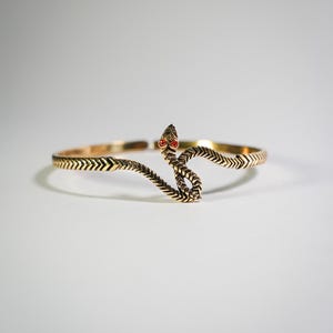 Op de afbeelding: Een goudkleurige metalen armband in de vorm van een kronkelende slang. De armband heeft een gestructureerd lichaam met een visgraatpatroon en twee kleine, ronde, felrode steentjes die als ogen in de kop van de slang zijn geplaatst. Het ontwerp is open.