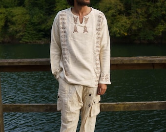May include: Cream-colored linen shirt and pants set. The long-sleeved shirt features intricate brown embroidery around the neckline, cuffs, and down the front. The pants have cargo pockets and button details. The outfit is worn on a wooden dock overlooking a lake.