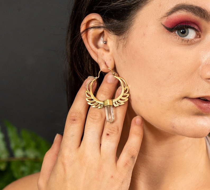 May include: Gold hoop earrings with a clear quartz crystal pendant. The earrings have a delicate, intricate design with a wing-like pattern.