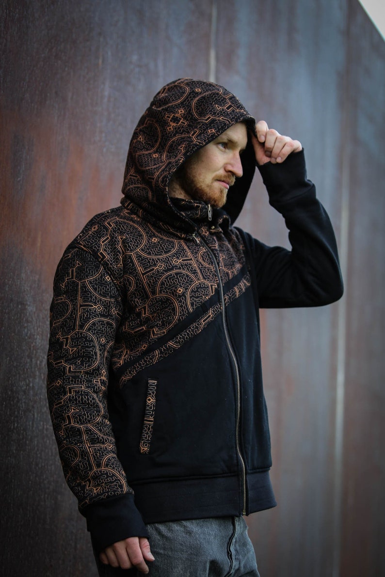 May include: A black zip-up hoodie with a brown geometric pattern. The hoodie has a hood with a drawstring and a zipper closure. The pattern is on the front and one sleeve of the hoodie.