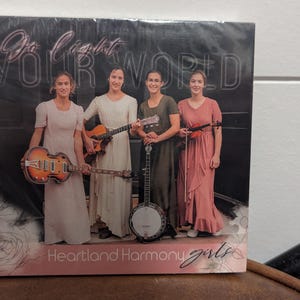 May include: CD cover featuring four women holding musical instruments. The text on the cover reads "Go Light Your World" and "Heartland Harmony Girls." The women are dressed in long dresses, with one holding a guitar, one a banjo, and one a violin.