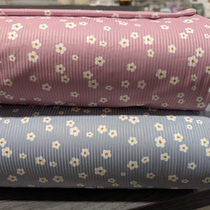 May include: Two rolls of fabric, one pink and one blue, are stacked on top of each other. Both fabrics have a ribbed texture and are patterned with small white flowers with yellow centers. The fabric is likely used for sewing or crafting projects.