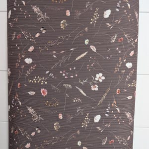 May include: A roll of dark brown ribbed fabric featuring a scattered print of delicate wildflowers, grasses, and small blossoms in muted shades of pink, white, beige, and tan. The material has a distinct horizontal texture throughout the surface.