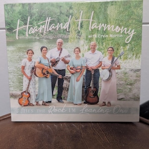 May include: Album cover for "Heartland Harmony" with Ervin Martin. The image features a group of people holding musical instruments, including guitars, a banjo, and a mandolin. The text "He's the Rock I'm Leaning On" is at the bottom.
