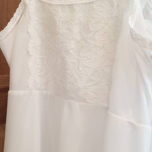 May include: White slip dress with delicate lace detailing on the bodice and straps. The dress is hanging on a white hanger against a wooden background. The lace has a floral pattern.