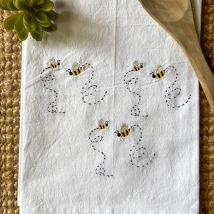 May include: White kitchen towel with embroidered bees flying in a circle. The bees are yellow and black with brown wings.