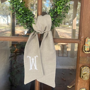 May include: A beige linen wreath bow with a white embroidered monogram "W" hanging on a wooden door with a green boxwood wreath.
