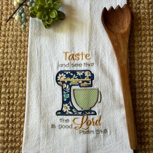 May include: White kitchen towel with an embroidered design of a blue and floral stand mixer and a green polka-dot bowl. The text reads "Taste and see that the Lord is good. Psalm 34:8". A wooden spoon rests on the side.