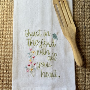 May include: White kitchen towel with embroidered text "Trust in the Lord with all your heart." Features a small bird and colorful floral accents. A wooden spatula rests on the towel, set against a textured background.