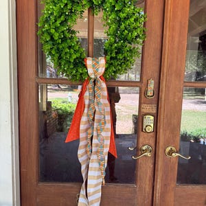 May include: A circular wreath made of vibrant green leaves, adorned with a decorative hanging bow. The bow features long, flowing ribbons in a combination of orange, white, and tan stripes, along with a patterned fabric in orange and teal.