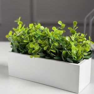 Könnte beinhalten: Ein rechteckiger weißer Pflanzkasten gefüllt mit leuchtend grüner künstlicher Vegetation. Die Blätter sind klein und rund und erzeugen ein dichtes, strukturiertes Aussehen. Der Pflanzkasten ist schlicht und modern, geeignet für die Inneneinrichtung.
