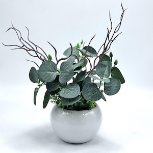 May include: An artificial eucalyptus plant in a white ceramic pot. The plant has round, grey-green leaves, small green buds, and dark brown branches. The pot is a simple, rounded shape, suitable for home decor.