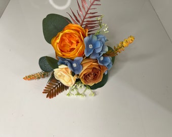 May include: A small bouquet of artificial flowers in shades of orange, brown, and blue. The bouquet includes roses, hydrangeas, and ferns.