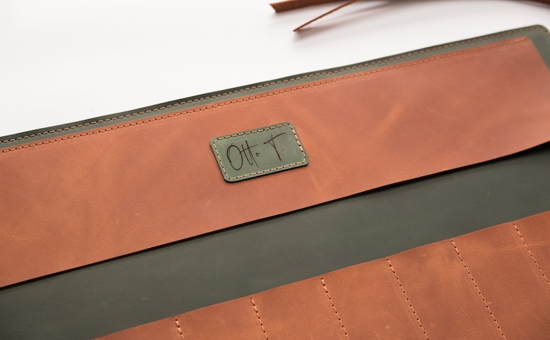 May include: A green leather tool roll with a brown leather flap. The flap has a small, rectangular leather patch with the text "Ott T." stitched in black.