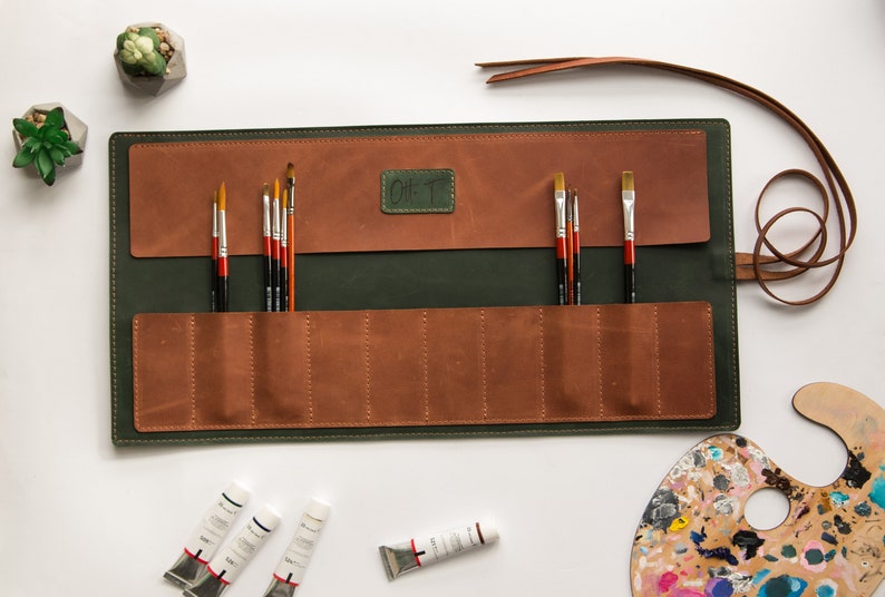 May include: A green leather roll-up case with brown leather pockets holds a set of paintbrushes. The case has a brown leather strap and a label that says "Gift".