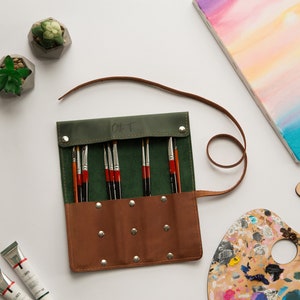 May include: A green and brown leather roll-up case with a strap, containing a set of paint brushes. The case has the word "Gift" written on it.