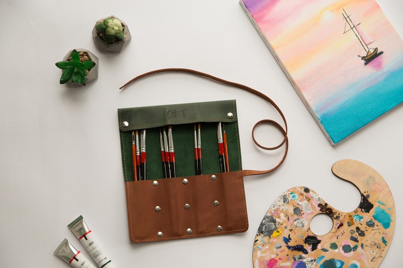 May include: A green and brown leather roll-up case with a strap, containing a set of paint brushes. The case has the word "Gift" written on it.