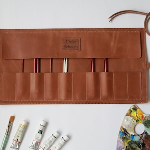 May include: A brown leather roll with eight slots for paintbrushes, a strap, and the name "Ulrike Selmons" embossed on the front. The roll is open and there are paintbrushes, paint tubes, and a paint palette on a white surface.