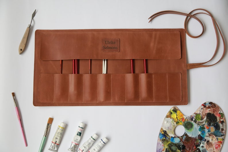 May include: A brown leather roll with eight slots for paintbrushes, a strap, and the name "Ulrike Selmons" embossed on the front. The roll is open and there are paintbrushes, paint tubes, and a paint palette on a white surface.