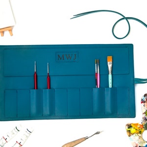 May include: Teal leather roll-up case with multiple pockets for holding paint brushes and tools. The case has a personalized monogram "MWJ" and a leather strap.