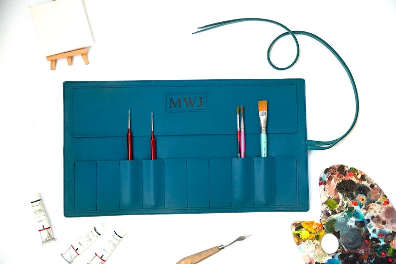 May include: Teal leather roll-up case with multiple pockets for holding paint brushes and tools. The case has a personalized monogram "MWJ" and a leather strap.