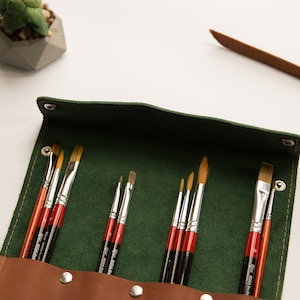 May include: A green leather roll-up case with a brown leather flap and snaps. The case is filled with a set of paint brushes with wooden handles and silver metal ferrules.