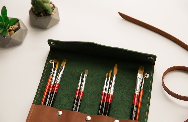 May include: A green leather roll-up case with a brown leather flap and snaps. The case is filled with a set of paint brushes with wooden handles and silver metal ferrules.
