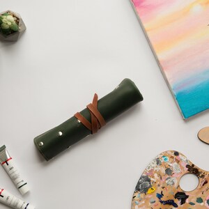 May include: A green leather roll-up case with a brown leather strap, two tubes of paint, a wooden paint palette with colorful paint, two small succulent plants in geometric concrete pots, and a canvas painting of a sailboat on a blue and pink sunset.