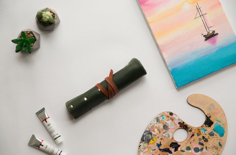 May include: A green leather roll-up case with a brown leather strap, two tubes of paint, a wooden paint palette with colorful paint, two small succulent plants in geometric concrete pots, and a canvas painting of a sailboat on a blue and pink sunset.