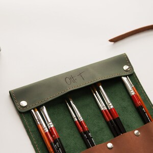May include: A green leather roll-up case with a brown leather strap and a snap closure. The case is filled with paint brushes and has the initials "O.H.T." embossed on the front.