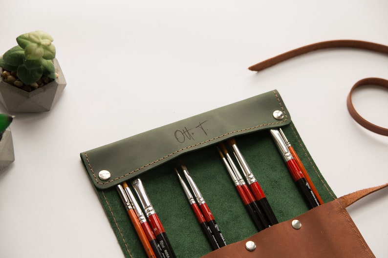 May include: A green leather roll-up case with a brown leather strap and a snap closure. The case is filled with paint brushes and has the initials "O.H.T." embossed on the front.