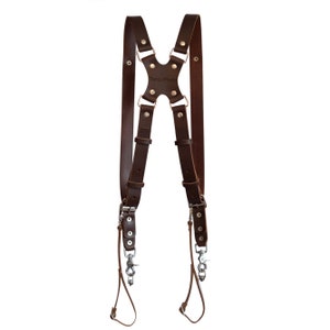 Può includere: Imbracatura per fotocamera in pelle marrone con hardware in metallo. L'imbracatura presenta cinghie regolabili e un design a croce sulla schiena. Il marchio "Tanzi Photo" è visibile. Progettato per fotocamere.