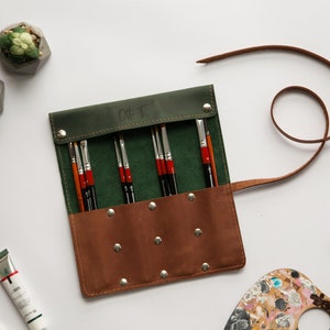 May include: A green and brown leather roll-up case with a strap, containing a set of paint brushes. The case has a label that says "Oh T".