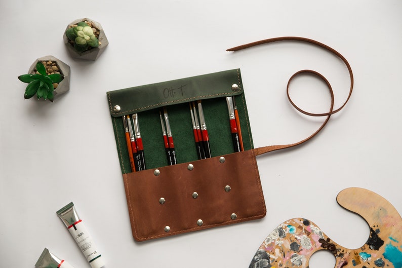 May include: A green and brown leather roll-up case with a strap, containing a set of paint brushes. The case has a label that says "Oh T".