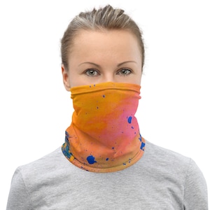 May include: A vibrant face covering with an abstract design. The neck gaiter features a blend of orange, pink, and blue paint splatters. The accessory is worn around the neck and covers the lower face.