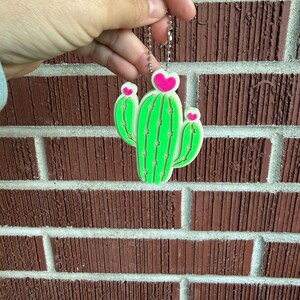 May include: A neon green cactus keychain with pink heart accents. The cactus design has a white outline and is held by a silver chain. The background is a red brick wall.