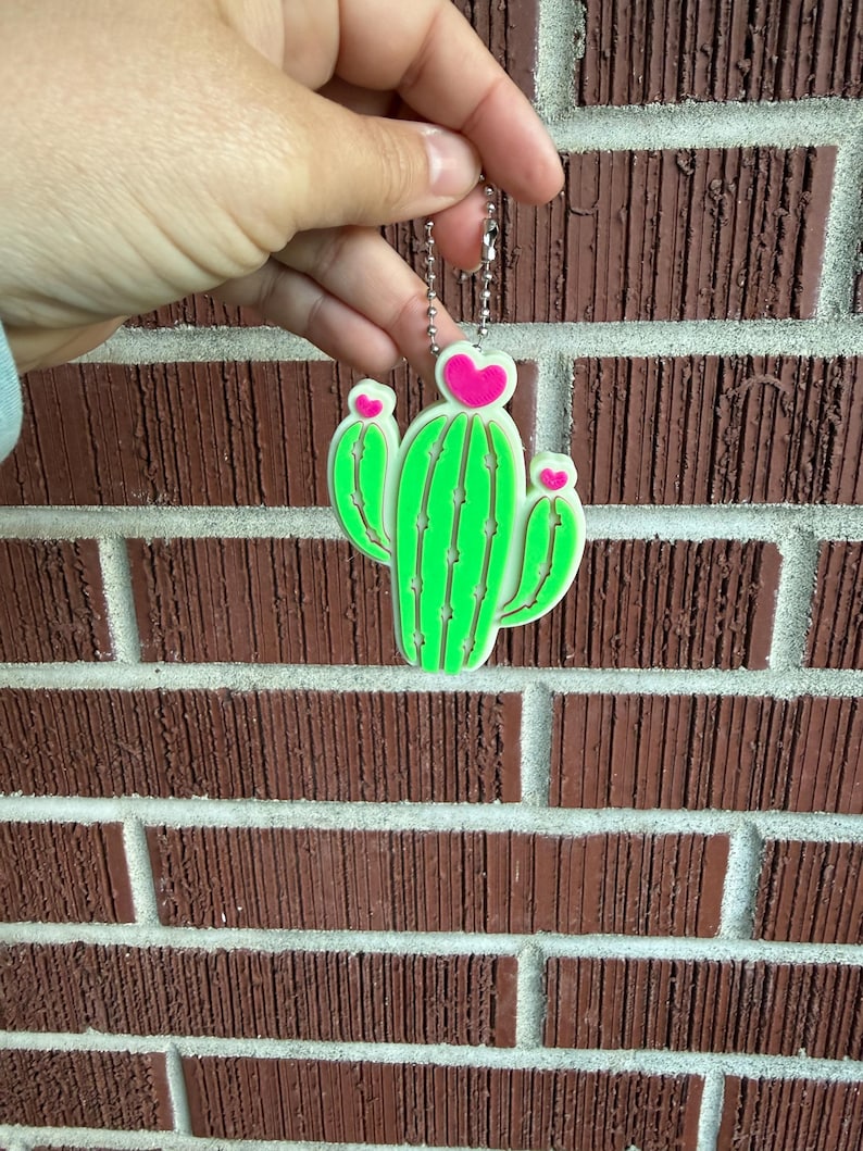 May include: A neon green cactus keychain with pink heart accents. The cactus design has a white outline and is held by a silver chain. The background is a red brick wall.