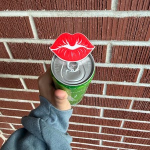 May include: A red and white lip-shaped can topper is placed on top of a green can. The can is held in front of a brick wall. The lip design has a white outline and is a fun accessory.