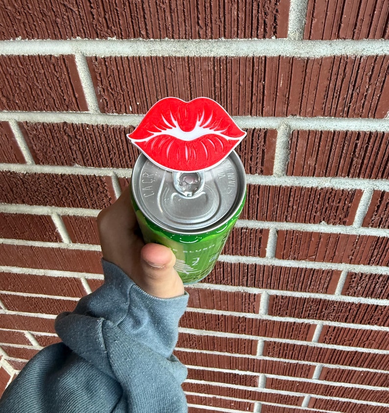 May include: A red and white lip-shaped can topper is placed on top of a green can. The can is held in front of a brick wall. The lip design has a white outline and is a fun accessory.