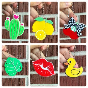 May include: A collection of colorful keychains featuring various designs. The keychains include a green cactus with pink hearts, yellow lemons, red cherries with a checkered bow, a green leaf, red lips, and a yellow duck.