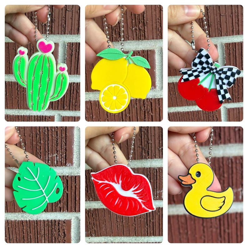 May include: A collection of colorful keychains featuring various designs. The keychains include a green cactus with pink hearts, yellow lemons, red cherries with a checkered bow, a green leaf, red lips, and a yellow duck.